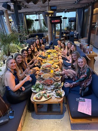 Large group of friends raising glasses for a celebratory dinner at a covered outdoor patio lounge, long table filled with shareable plates, salads, fries and cocktails under cozy lights and heaters — lively girls’ night out vibe.
