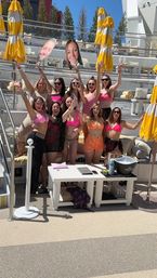 Group of friends cheering at a sunny outdoor pool lounge with yellow-and-white umbrellas, women in pink swimsuits and bright summer outfits holding oversized face cutouts and posing around a white cabana table with drinks.