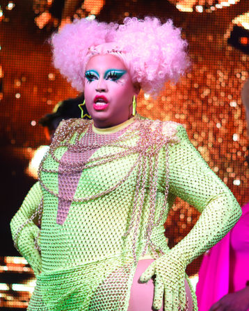 Neon-glam drag performer on a glittering club stage wearing a lime mesh studysuit with chain accents, fluffy pink afro wig, bold teal eye makeup, star earrings and vibrant pink lips.