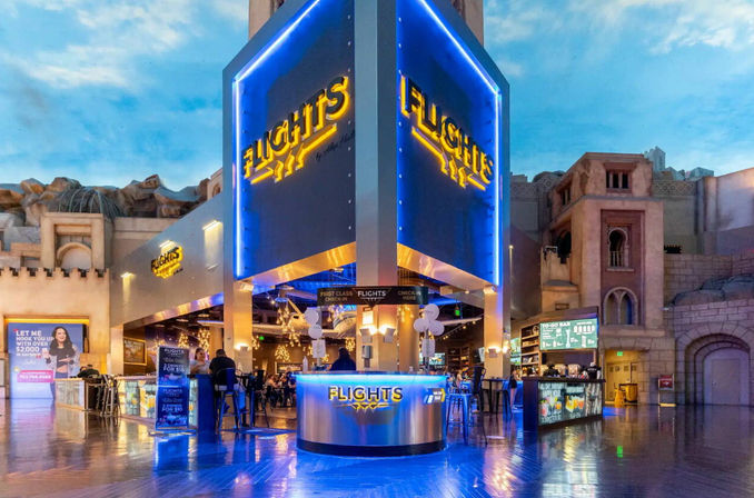 Vibrant blue-lit, aviation-themed indoor bar and lounge with a large illuminated central kiosk, patrons at counters, chandeliers overhead and glossy floor reflections inside an entertainment complex.