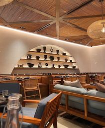 Cozy modern restaurant interior with woven wood ceiling and exposed beams, arched wall shelves filled with pottery, rattan chairs and cushioned wooden seating under warm ambient lighting.