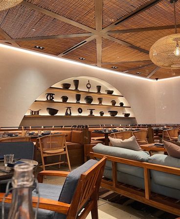Cozy modern restaurant interior with woven wood ceiling and exposed beams, arched wall shelves filled with pottery, rattan chairs and cushioned wooden seating under warm ambient lighting.