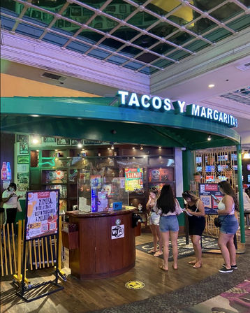 Indoor mall Mexican restaurant entrance with green awning and bright "TACOS Y MARGARITAS" neon sign, hostess podium and menu displays, and a small group of customers waiting and checking phones.