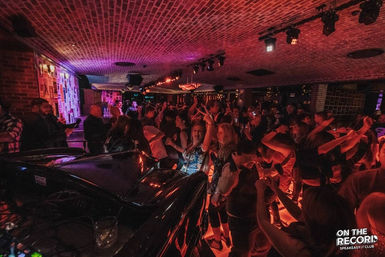 Energetic speakeasy-style nightclub interior with a brick-vaulted ceiling, packed dance floor, DJ booth in the foreground and colorful red-purple lighting as people dance and hold drinks.
