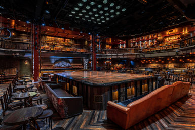 Empty cabaret-style theater with a raised square wooden stage at center, leather sofas and round wooden tables around it, two-level balcony seating, warm amber lighting and dark herringbone wood floor