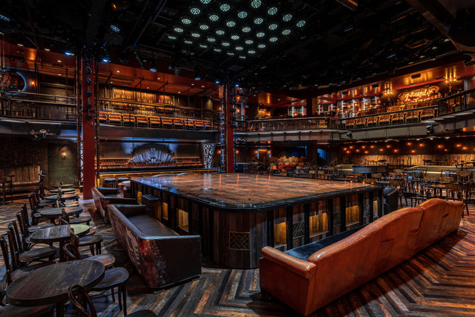 Empty cabaret-style theater with a raised square wooden stage at center, leather sofas and round wooden tables around it, two-level balcony seating, warm amber lighting and dark herringbone wood floor