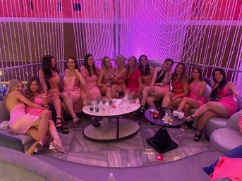 Large group of women in pink dresses seated on a curved sofa in a modern lounge with pink-purple lighting and a beaded curtain backdrop, cocktails on low tables — lively girls' night out.