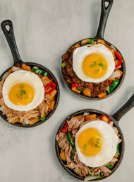Three mini cast-iron skillet brunch hashes — diced potatoes, peppers and shredded meat, each crowned with a sunny-side-up egg and chopped green onions on a light tabletop.