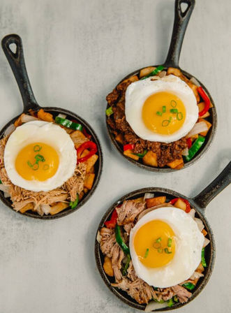 Three mini cast-iron skillet brunch hashes — diced potatoes, peppers and shredded meat, each crowned with a sunny-side-up egg and chopped green onions on a light tabletop.