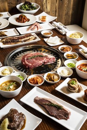 Korean BBQ spread with a marbled ribeye on a tabletop grill topped with rosemary, surrounded by colorful banchan, dipping sauces, soups, rice, and raw cuts on white plates on a wooden table.