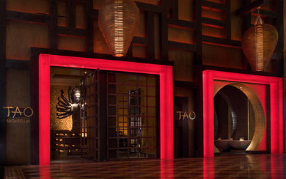 Red-lit upscale Asian-inspired nightclub entrance with a dramatic multi-armed statue peeking through lattice doors, a circular arch alcove, and woven pendant lanterns.