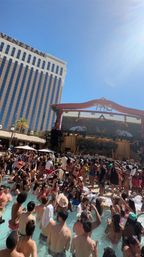 Sunny Las Vegas resort pool party with hundreds of people in swimsuits packed in and around a crowded pool, live DJ on a decorated stage and a tall hotel tower rising under a clear blue sky.