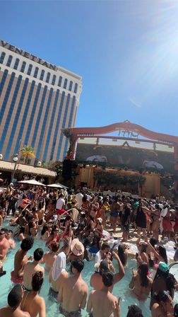 Sunny Las Vegas resort pool party with hundreds of people in swimsuits packed in and around a crowded pool, live DJ on a decorated stage and a tall hotel tower rising under a clear blue sky.