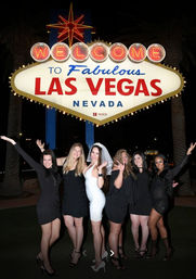 Six women, one in a white bridal dress and veil, posing at night beneath the illuminated 'Welcome to Fabulous Las Vegas Nevada' sign — playful bachelorette party photo