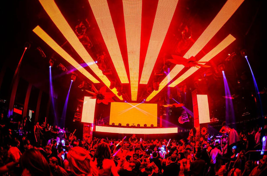 Energetic nightclub scene: packed dance floor bathed in red-orange LED beams, DJ booth with large screens overhead, purple spotlights and a dancing crowd waving glow sticks.