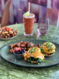 Bright brunch scene: two eggs Benedict with wilted spinach and gooey hollandaise, crispy home fries, a cup of mixed berries, and a salted‑rim Bloody Mary on a green marble table.