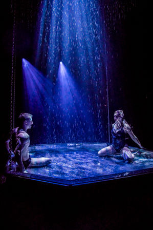 Two performers in a shallow stage pool beneath cascading water and vibrant blue spotlights, a theatrical aquatic dance duet on a dark stage.