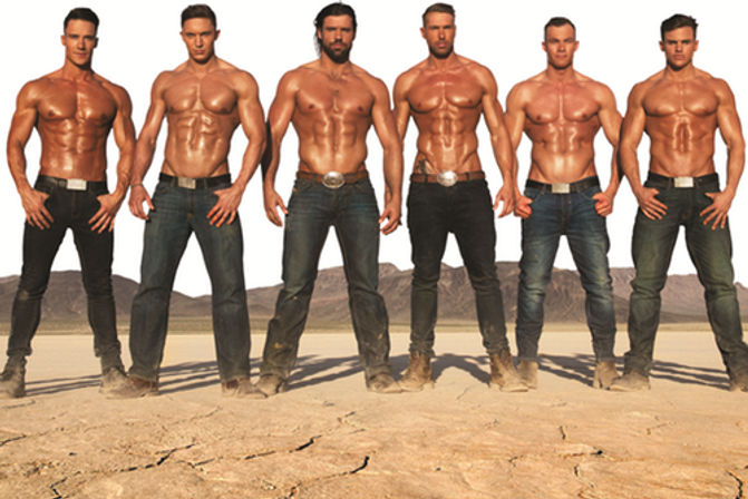 Six shirtless muscular male models in jeans and boots lined up on a cracked dry desert lake bed with mountains in the background — outdoor fitness photoshoot