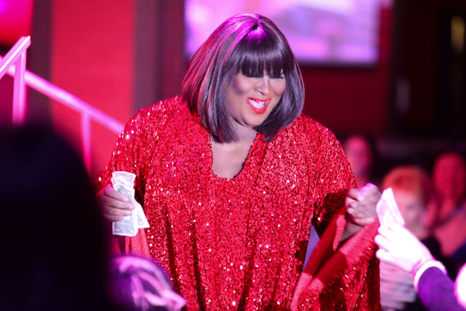 Glamorous performer in a sparkling red sequined gown and bob wig smiling as they accept dollar bills from the audience during a lively nightclub cabaret show.