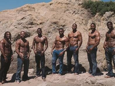 Seven shirtless, muscular men in jeans posing on a sunlit sandy beach in front of a rocky cliff.