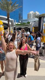 Sunny rooftop pool party with palm trees and high-rise buildings; excited woman in a beige mesh dress raises her arms while friends in colorful swimwear mingle under yellow umbrellas.