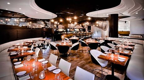 Modern upscale restaurant interior with curved white banquettes, polished wood floors, wooden dining tables set with wine glasses and candles, black-and-white swivel chairs, ambient pendant lighting and a central bar.