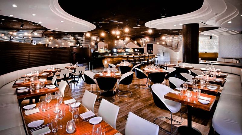 Modern upscale restaurant interior with curved white banquettes, polished wood floors, wooden dining tables set with wine glasses and candles, black-and-white swivel chairs, ambient pendant lighting and a central bar.