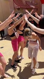 Bachelorette party on a sunny resort pool deck: women in black bikini tops and pink wraps form a cheering human arch while the bride-to-be in white lace outfit, sash and sunglasses ducks through.