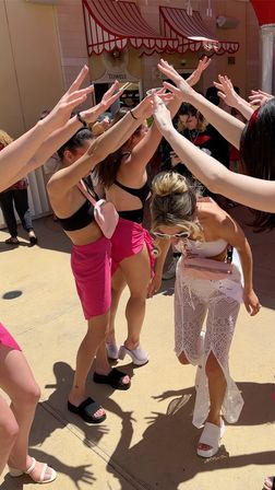 Bachelorette party on a sunny resort pool deck: women in black bikini tops and pink wraps form a cheering human arch while the bride-to-be in white lace outfit, sash and sunglasses ducks through.