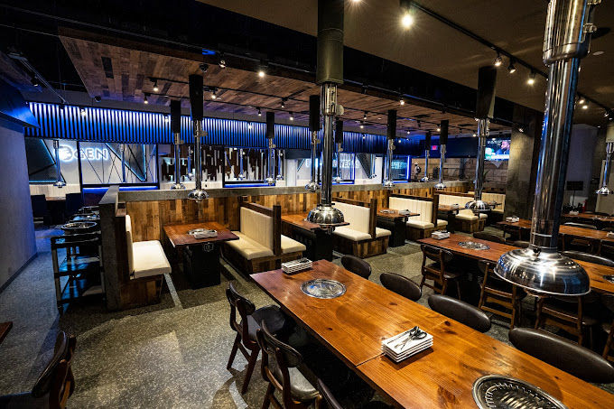 Spacious modern Korean BBQ restaurant interior with wooden booths, long communal tables, built-in tabletop grills and chrome vent hoods under warm wood and blue accent lighting.