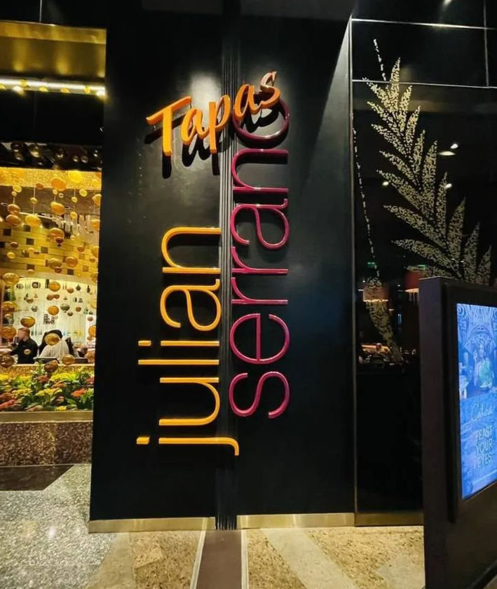 Sleek black restaurant entrance with glowing orange “Tapas” sign and tall pink-orange vertical lettering, glass window showing hanging gold orbs and a fresh produce display.