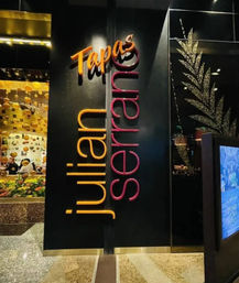 Sleek black restaurant entrance with glowing orange “Tapas” sign and tall pink-orange vertical lettering, glass window showing hanging gold orbs and a fresh produce display.