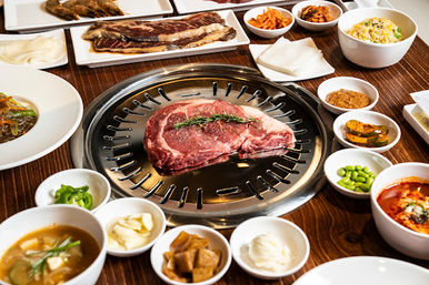 Tabletop Korean BBQ grill with a raw ribeye steak topped with rosemary, surrounded by assorted banchan, rice, soup, and dipping sauces on a wooden table