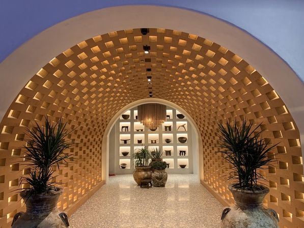 Warm honeycomb-patterned arched corridor with illuminated square niches displaying pottery, central chandelier, and large potted plants in rustic urns.
