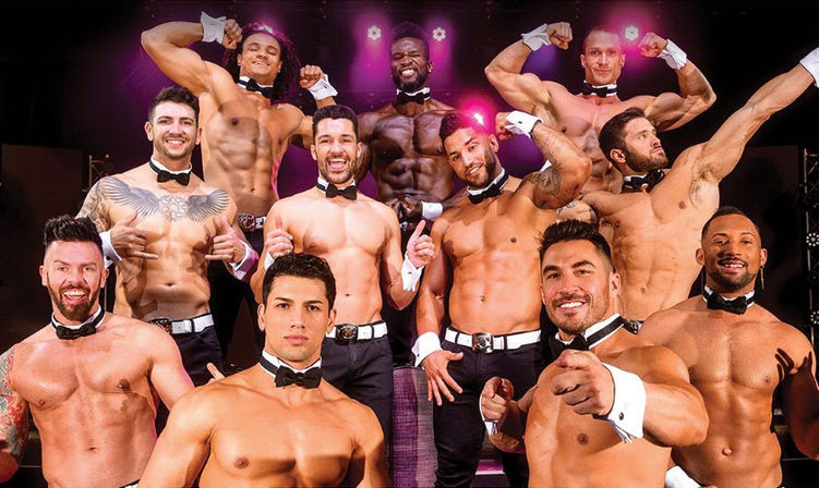 Group of shirtless male revue dancers wearing bow ties and cuffs, flexing and smiling on a lit stage with purple spotlights