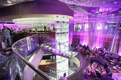 Glamorous multi-level nightclub lounge interior with purple ambient lighting, illuminated glass column and spiral staircase, upper bar area and groups of people socializing on curved lounge seating.