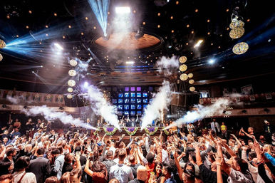 Packed nightclub dance floor with hands-up crowd facing a DJ booth, colorful stage lights, smoke cannons and hanging disco balls.