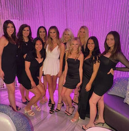 Group of eleven women for a girls’ night out, one in a white mini dress surrounded by friends in black cocktail dresses, posing and smiling in an upscale lounge with purple-pink beaded curtain and ambient lighting.