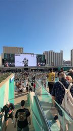 Sunny rooftop resort pool deck packed with people watching a hockey game on a giant outdoor video screen, fans in swimwear and a yellow hockey jersey, glass railings and high-rise hotels in the background.
