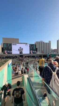 Sunny rooftop resort pool deck packed with people watching a hockey game on a giant outdoor video screen, fans in swimwear and a yellow hockey jersey, glass railings and high-rise hotels in the background.