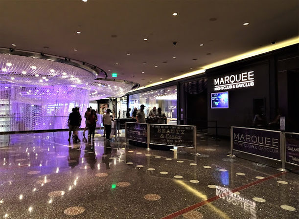 People walking through a glossy casino corridor with a large purple-lit crystal chandelier installation, reflective terrazzo floor, and rope-barrier nightclub entrance with illuminated signage.