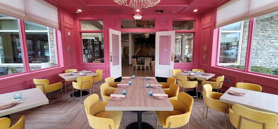 Playful pink dining room interior with wooden tables, mustard-yellow velvet chairs, large windows and a central chandelier in a sunlit café