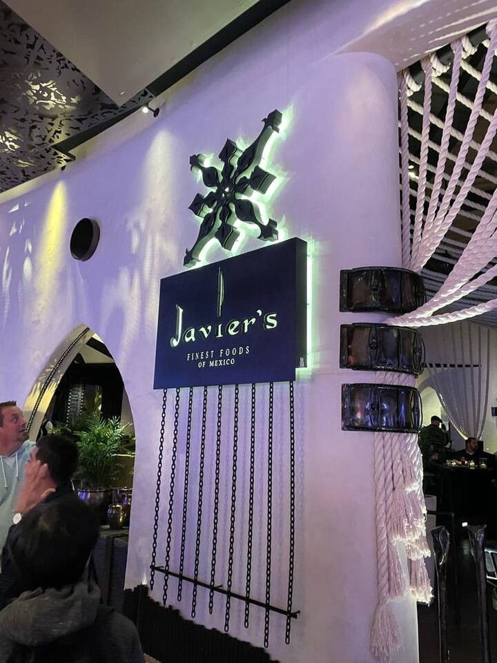 Upscale Mexican restaurant interior with glowing logo on a curved white wall, dangling chains and thick braided ropes, purple mood lighting and diners in the foreground.