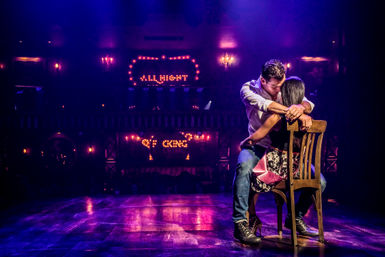 Two performers in an intimate embrace on a wooden chair center stage under purple-pink spotlights with glowing marquee lights in a dim cabaret-style theater.