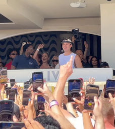 Daytime crowd holding up smartphones to film a smiling performer in a visor and sleeveless shirt on an elevated day-club stage, with security and photographers behind him.