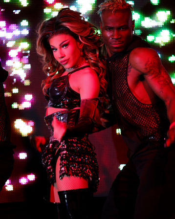High-energy nightclub stage scene: glamorous performer in a sequined black crop top, fringed mini skirt, fishnet stockings and thigh-high boots with dramatic makeup and big wavy hair poses with a muscular backup dancer in a mesh top against colorful LED lights.
