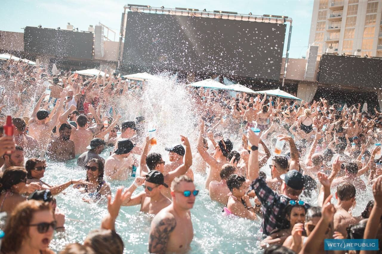 Crowded daytime outdoor pool party with people splashing, cheering, and raising drinks under bright sun and umbrellas