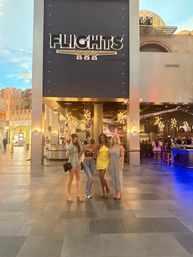 Four friends posing under a large illuminated entrance sign inside a lively indoor dining and entertainment complex with chandelier-style pendant lights and wide tiled floor