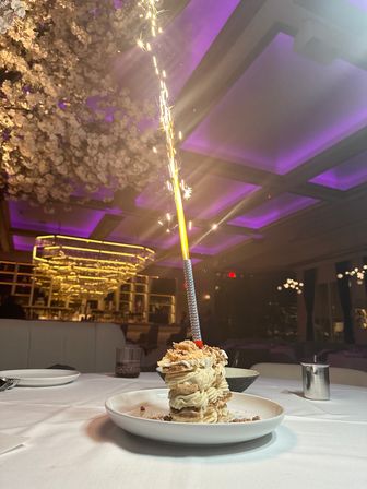Celebratory layered cream pastry on a white plate topped with a lit sparkler, set on a white tablecloth in an upscale restaurant interior with purple ceiling lighting and hanging floral decor.