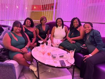 Six friends in stylish evening wear smiling around a cocktail table in a pink‑lit lounge with plush seating and beaded curtain backdrop — girls’ night out, cocktail lounge nightlife